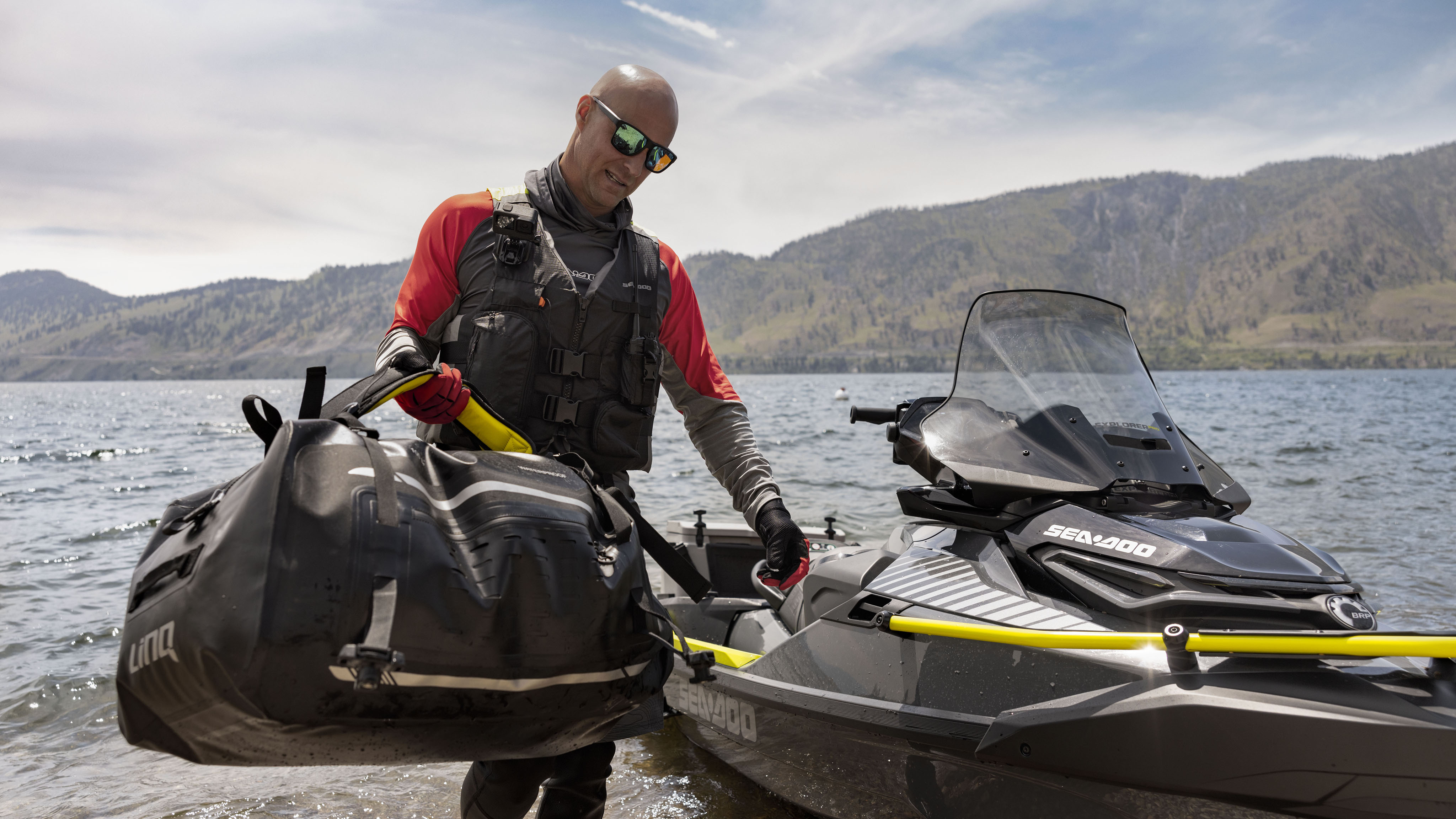一位男士提着 Explorer 防水包，身旁是一台 Sea-Doo Explorer Pro 摩托艇